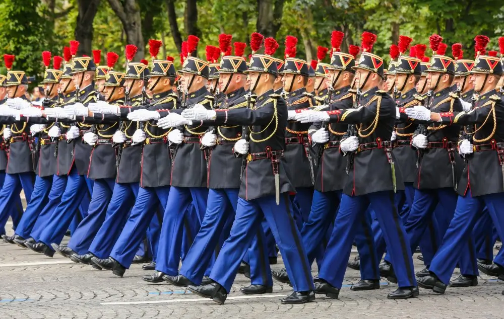 Défilé militaire de soldats français en uniforme de cérémonie avec plumets rouges et gants blancs.