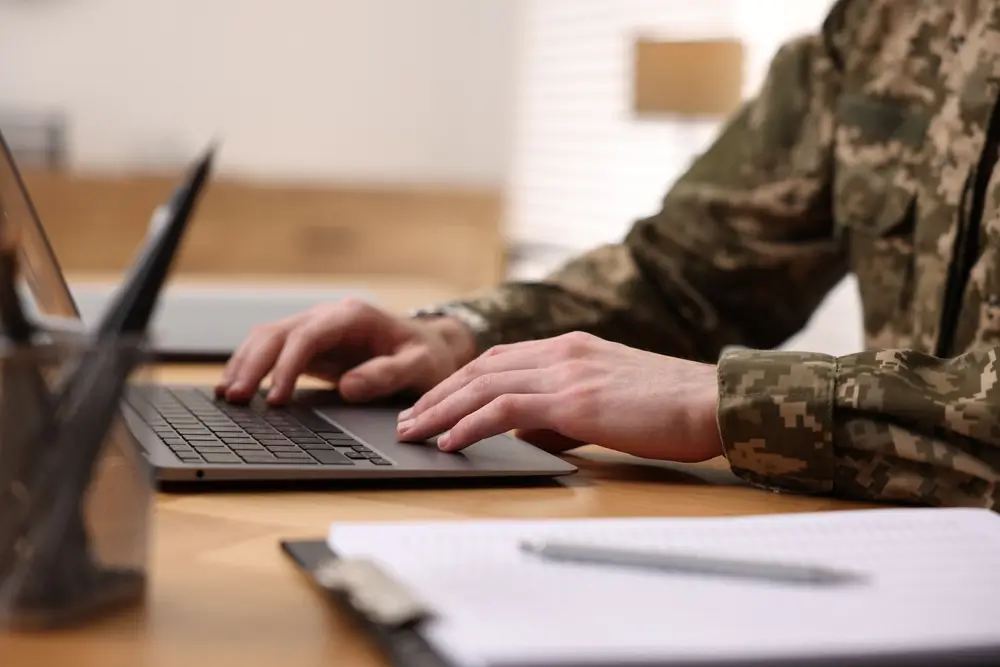 Militaire en treillis travaillant sur un ordinateur pour préparer son concours, avec un dossier à ses côtés.